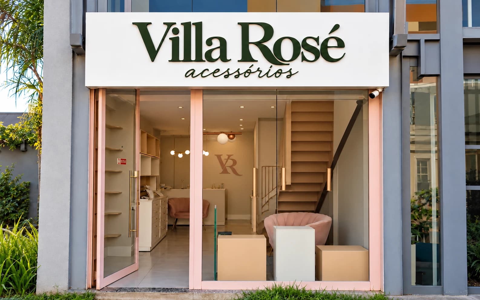 Fachada da Villa Rosé acessórios, com o nome em verde sobre painel branco, esquadrias rosé e o monograma VR ao fundo da loja.
